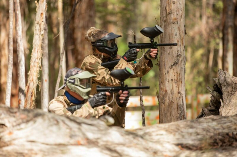 Two players aim paintball markers from behind trees in the woods
