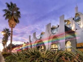 A futuristic looking building surrounded by palm trees and greenery. A dash of rainbow in front.