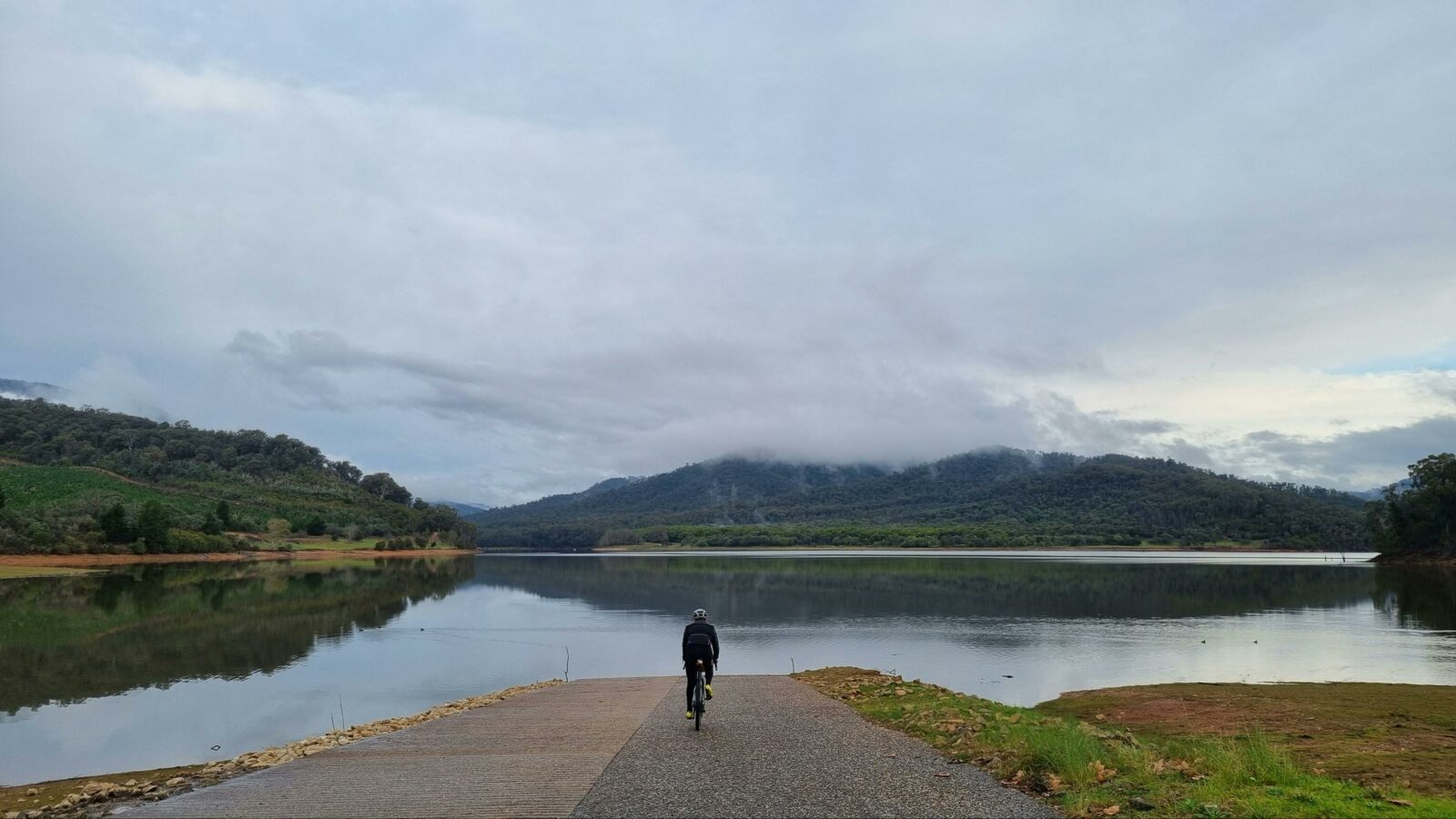 Cyclist on ramp, lake, hills with grass and trees, shadows in water, grey clouds, low cloud on hills