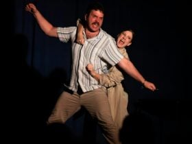 A man with arms outstretched, held around the torso by a woman in a trenchcoat. Both are singing.