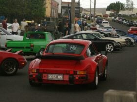 red classic Porsche driving past rows of cars at the marque collective cars and coffee