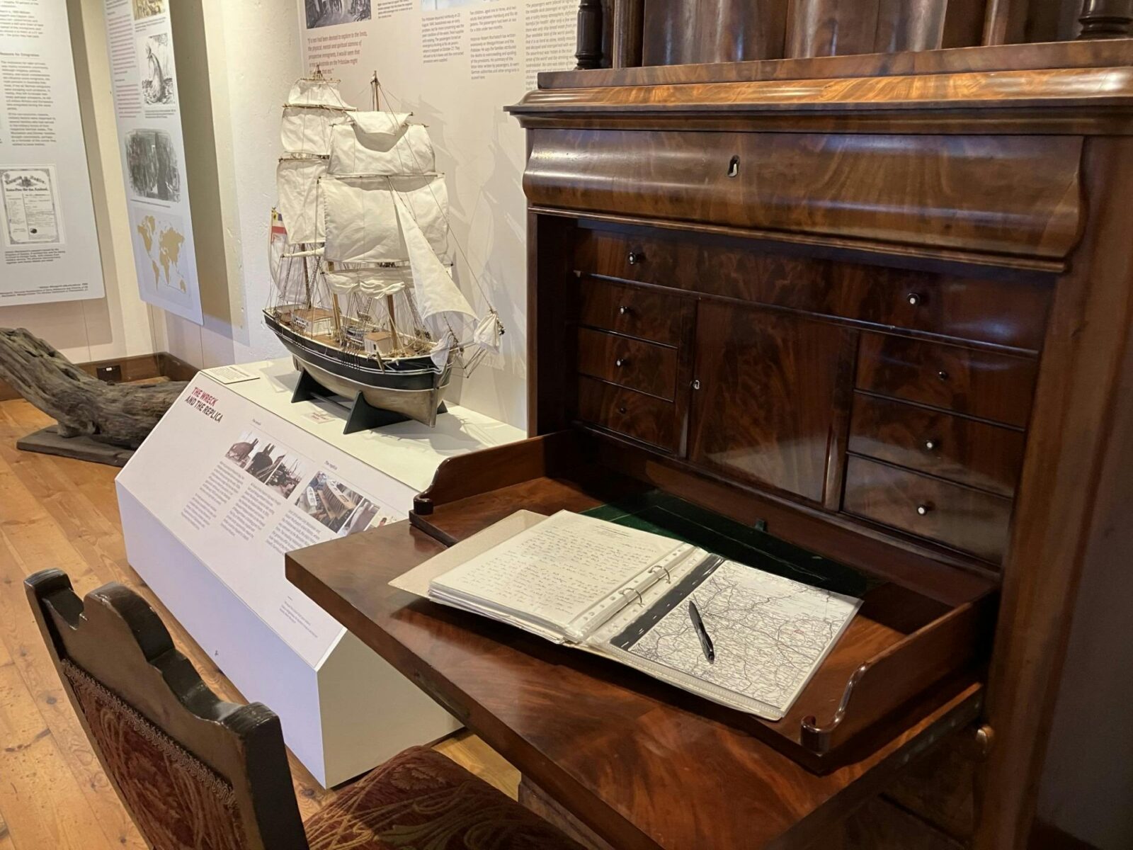Historic desk with open journal beside a model sailing ship in a museum display.