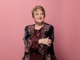 Comedian Denise Scott in front of a pink background, smiling