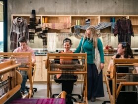 Three women seated at looms with instructor standing between them inside workshop