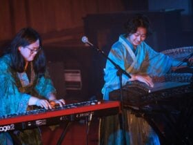Two musicians in robes perform on guzheng and keyboard under stage lights, smiling as they play.
