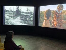 Person seated in a dark gallery watching two large video screens, one black-and-white, one in color.
