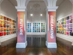 Two pillars stand in the centre of a gallery space with hundreds of colourful 30x30cm paintings hung