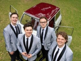 Four band members standing in front of a car with its doors open, parked on grass.