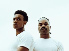 Two men in white t-shirst look at us while standing in front of a white background