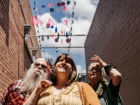 3 people look up, interested in th hanging artworks in a laneway, cloudy blue sky in the background.