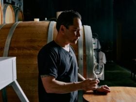 Winemaker in black teeshirt assessing wine in a glass, barrels in background