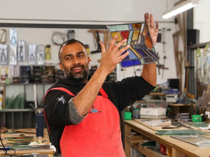 A male student with a darker complexion in a red apron and black shirt holding up his leadlight pane