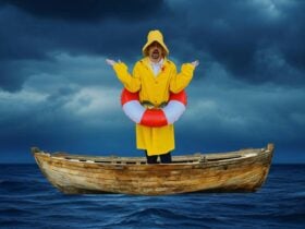 Jeremy Moses stands with arms up in a confused pose, in an old wooden boat in the ocean.