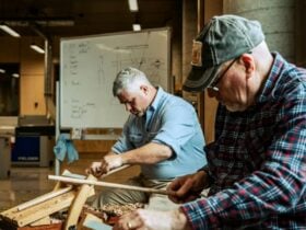 Two men seated and carving wood inside workshop