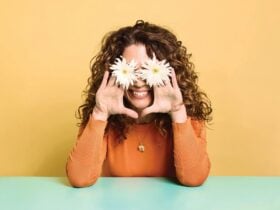 Kitty Flanagan holding two flowers over her eyes, smiling with her elbows leaning on a desk