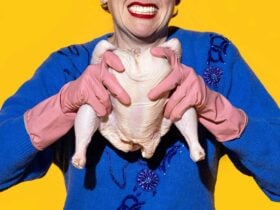 A woman in blue with pink rubber gloves and bright red lips holds a raw chicken and grimaces.