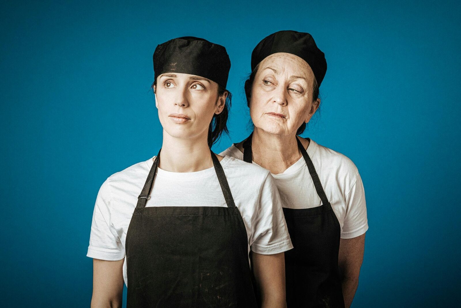 Young woman stares dreamily into the distance. Old woman scrutinises her. Both are deli workers