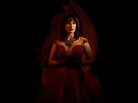 A striking, red-headed woman stands is a flowing red dress against a black background.