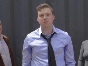 Two men and a woman stand in front of a grey wall. The 1st man and the woman look at the central man