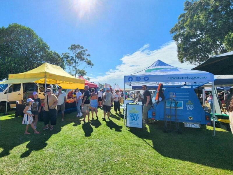 Market stalls on green grass with the sun shining and people milling around shopping