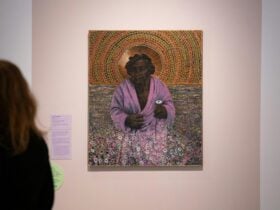 A painting of an elderly Australian Aboriginal woman surrounded by purple flowers hands on a wall.