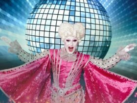 A performer in dramatic makeup and flamboyant pink costume in front of a giant disco ball