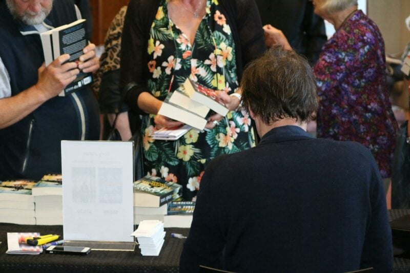 A man (an author) is seated with his back to camera, two women wait for him to sign their books
