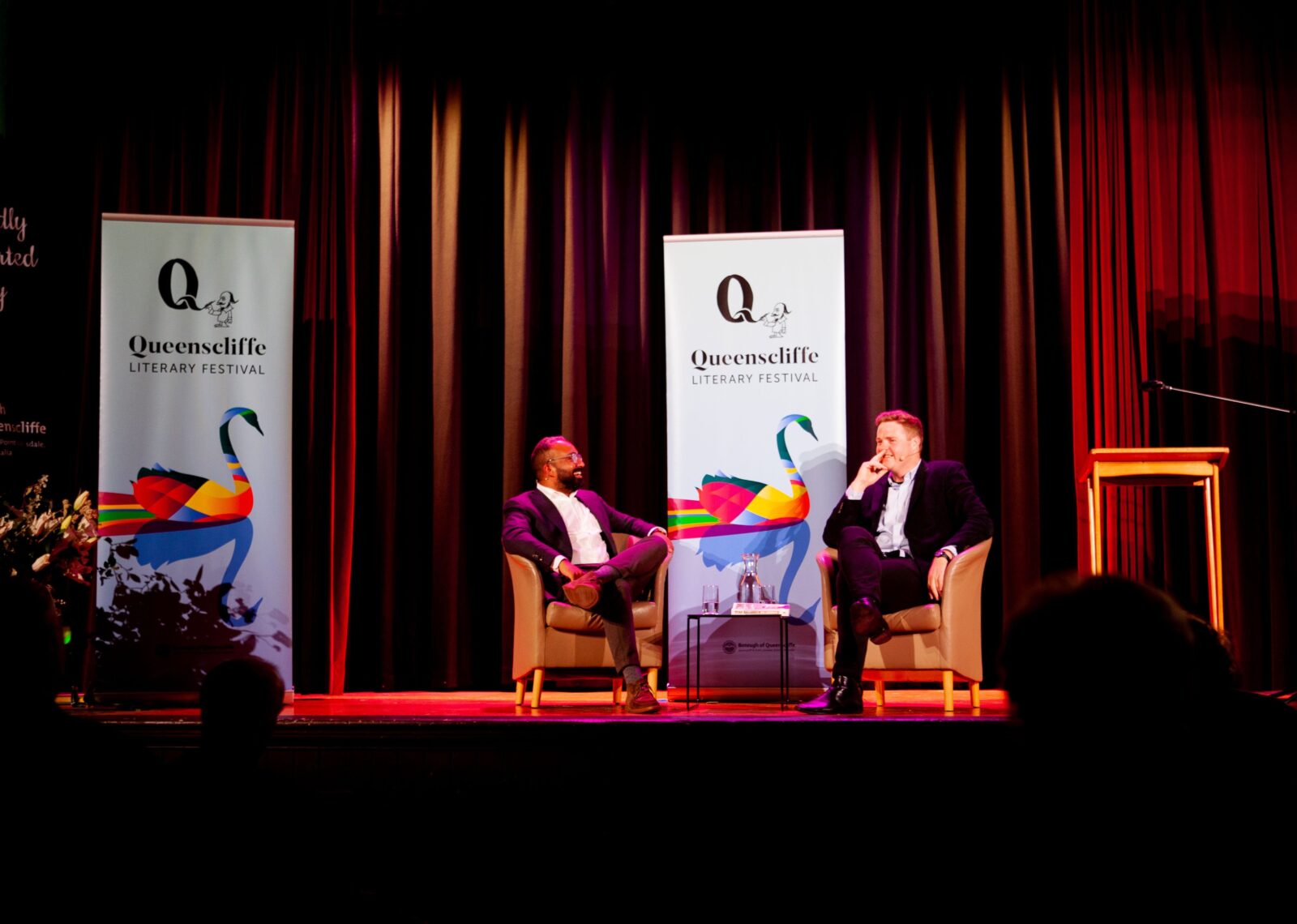 Two men sit on stage; there are QLF banners behind them