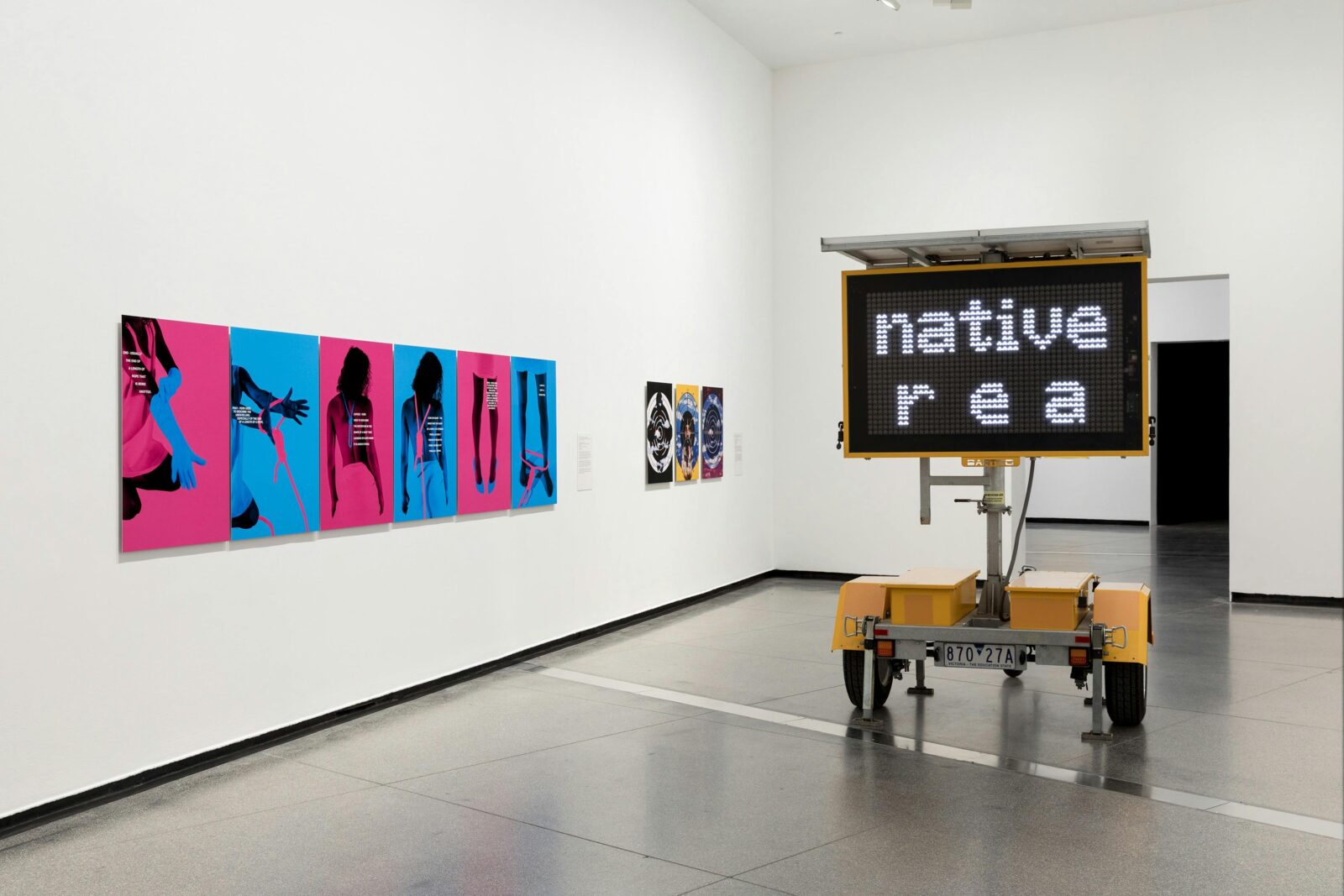 A view of a room in a gallery space. Brightly coloured artworks are shown, including a road sign