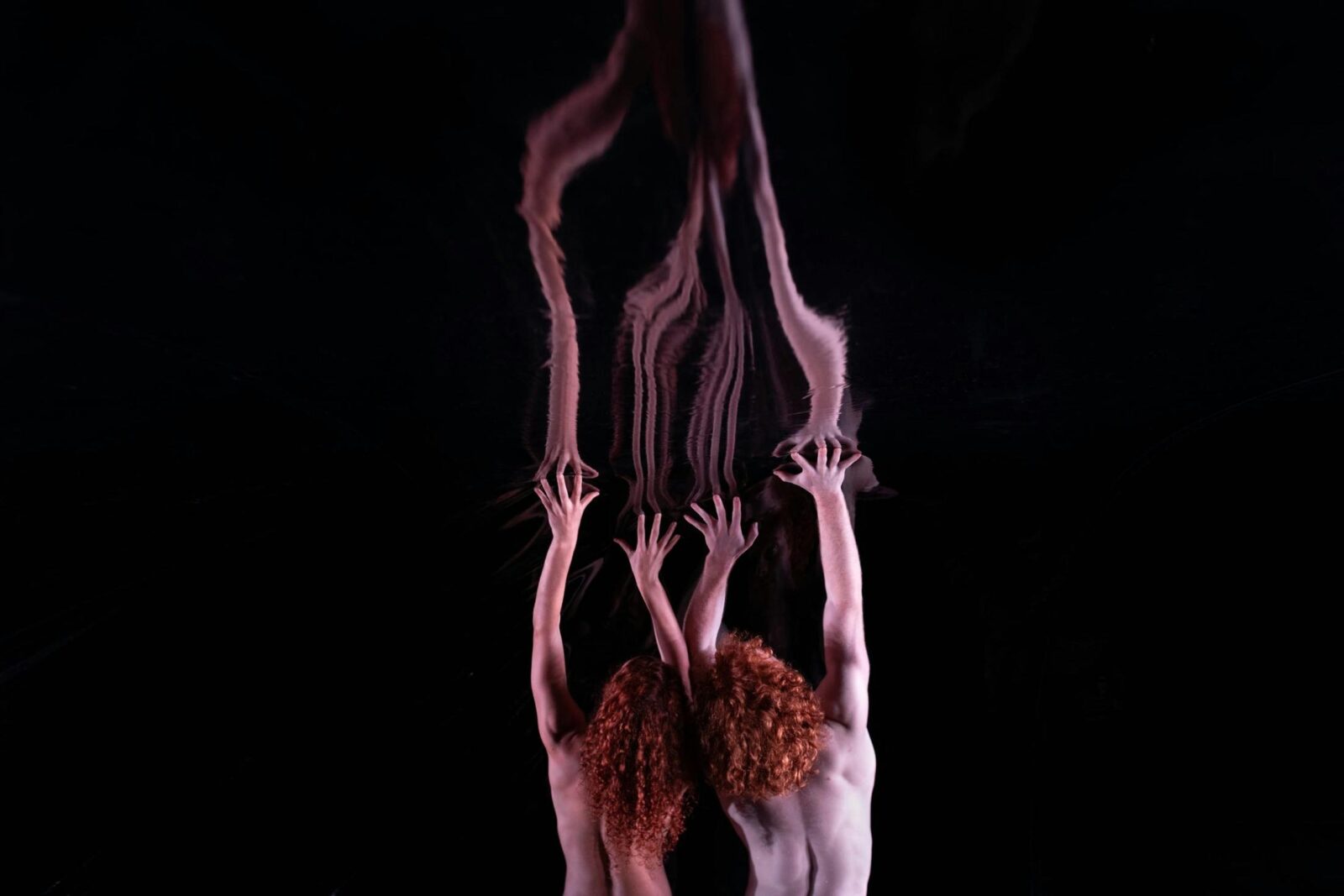 Two people with red hair reach both arms up towards a reflective surface. The background is black.