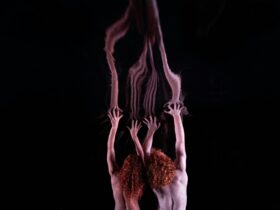 Two people with red hair reach both arms up towards a reflective surface. The background is black.
