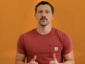 White man in red Carhartt t-shirt giving two thumbs up against an orange wall background.