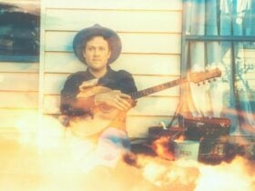 Man playing guitar on a porch with a window and plants in the background