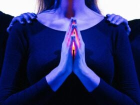 The torso of a woman bathed in blue light, glowing hands pressed in prayer, 2 hands on her shoulders