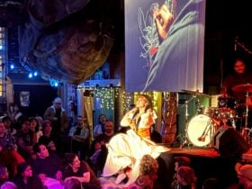 Photo of a performer in a large hall with a video screen and drummer on stage