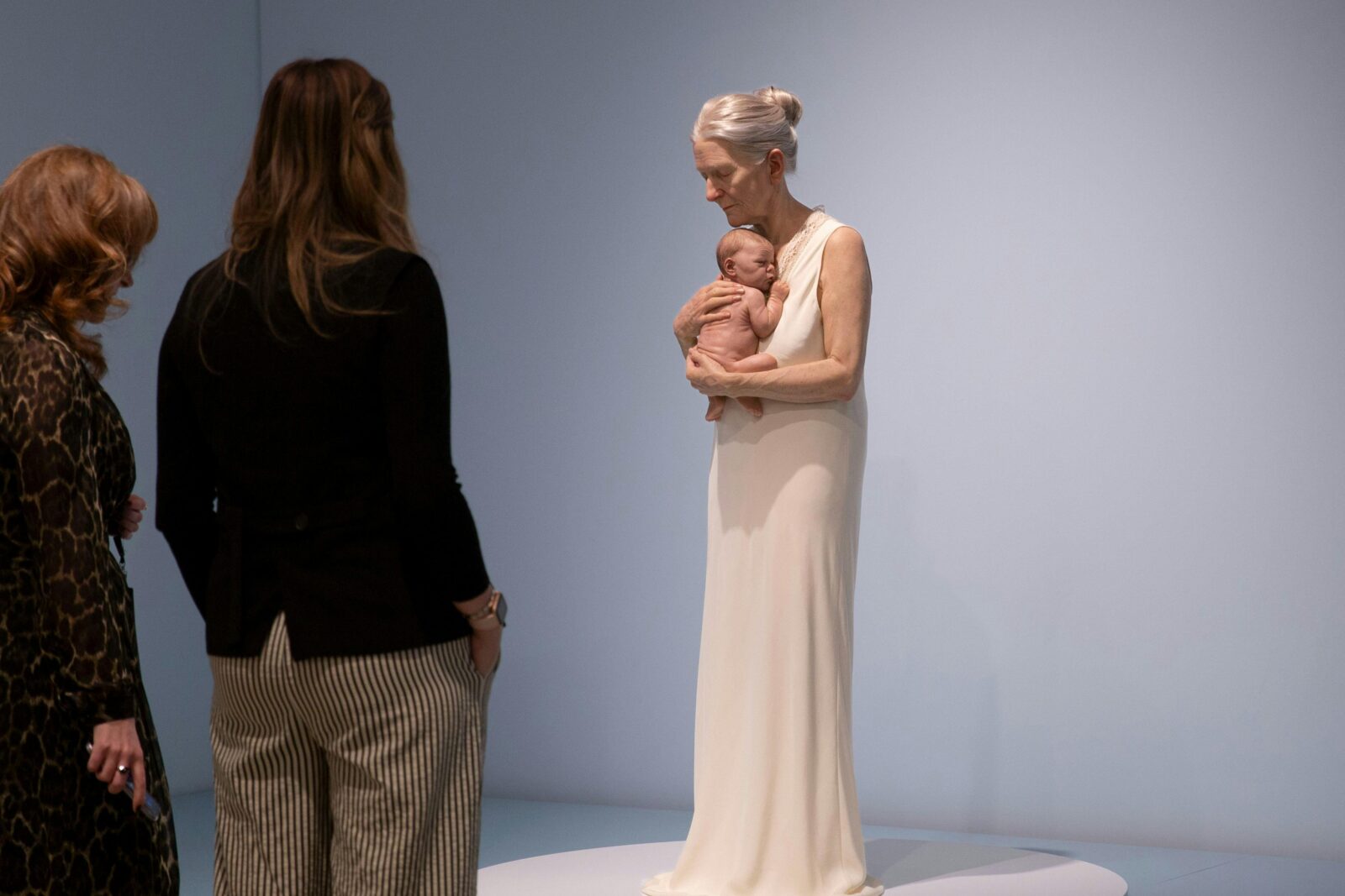 Two women view a hyperrealistic sculpture of an elderly woman in a nightgown holding a baby