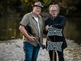 Two men, one holding a mandolin, the other a lagerphone, standing in front of a river.