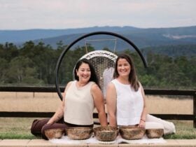 Two women seated outdoors with singing bowls and forest views behind them.