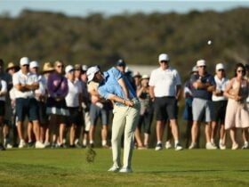 A golfer hits an approach shot with a large crowd watching in the late afternoon sun.