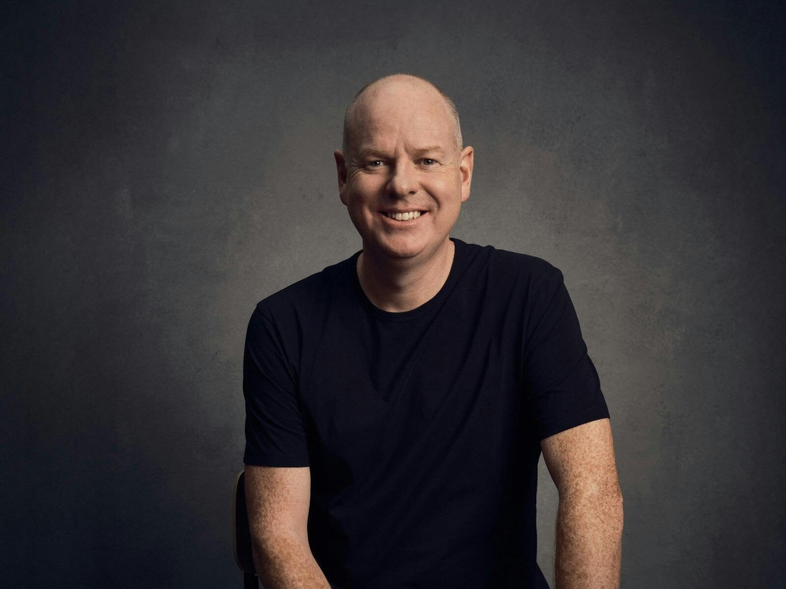 Comedian Tom Gleeson sitting on a stool, smiling