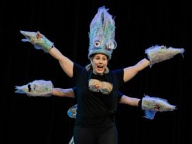 Performer in colorful squid-like costume with multiple arms made of trash, extended on a dark stage.
