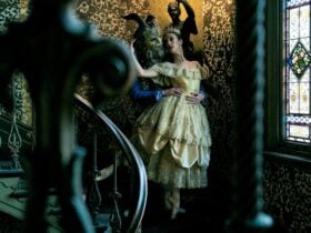 Dancer in a gold gown with a costumed partner on an ornate staircase.