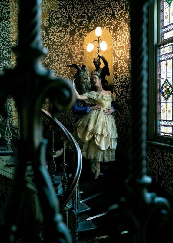 Dancer in a gold gown with a costumed partner on an ornate staircase.