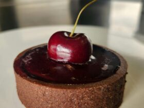 A chocolate tart sits on a white plate, it has a cherry jelly filling with a fresh cherry on top