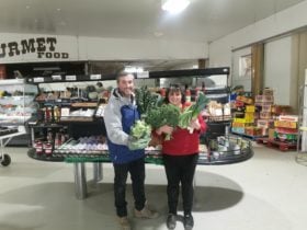 Two people holding fresh vegetables behind them is two counters with produce for sale