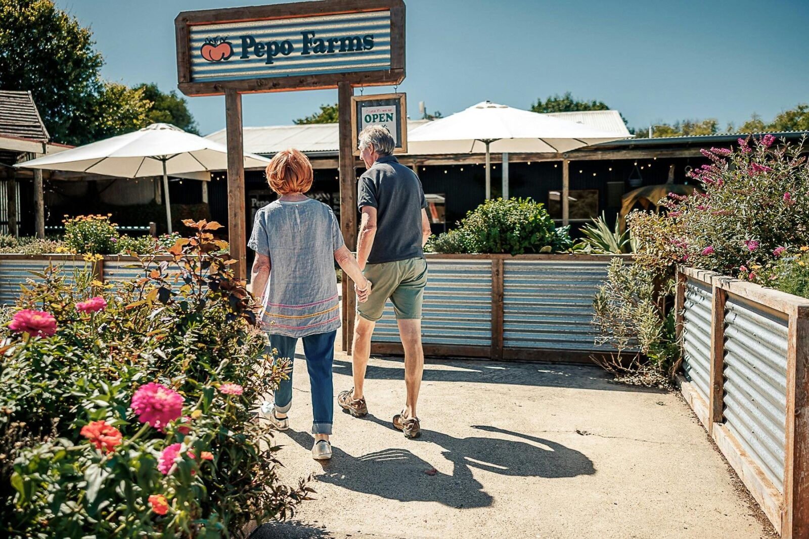 Couple walks hand-in-hand through garden toward Pepo Farms shopfront on a sunny day.