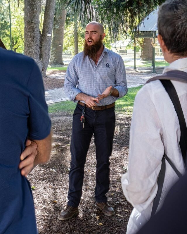Tour guide speaking to two visitors in the Town Hall Gardens. Large tree trunks in the background.