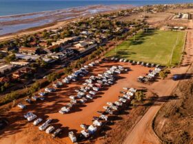 Aerial view of RV overflow with rows of caravans on red dirt beside sports field and coast