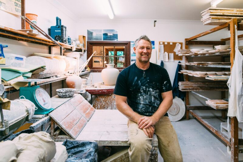 This photo shows Ian sitting in his pottery studio. There is a wheel thrown pot on the wheel.