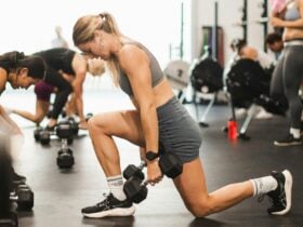 A woman in a grey workout set performing a weighted lunge inside a gym.